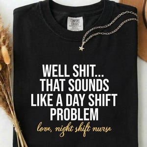 May include: Black t-shirt with the text "WELL SHIT... THAT SOUNDS LIKE A DAY SHIFT PROBLEM love, night shift nurse" in white and gold lettering. A silver necklace with a star pendant is on the shirt.