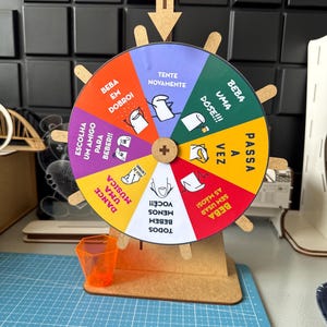 May include: A colorful wheel game with various instructions in Portuguese. The wheel is mounted on a wooden stand with an orange shot glass. The wheel has segments with phrases like "Try Again" and "Drink a Dose!!!".