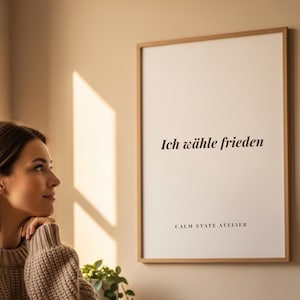 May include: A framed print with the text "Ich wähle frieden" in black, displayed on a beige wall. The print is in a light brown wooden frame. The words "CALM STATE ATELIER" are printed below the main text. A woman is looking at the print.