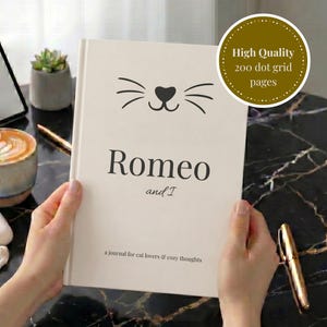 Puede incluir: Un diario de tapa dura color crema con un gráfico negro de nariz y bigotes de gato. El texto dice Romeo and I, a journal for cat lovers & cozy thoughts. Una insignia dorada circular indica 200 dot grid pages.
