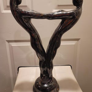 Op de afbeelding: Een donkere, glanzende sculptuur die twee verweven figuren afbeeldt, die een driehoekige vorm vormen. De figuren zijn bij de handen verbonden, met een afgeronde basis. De sculptuur heeft een reflecterend oppervlak met rode en gouden accenten.