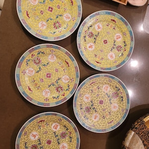Peut inclure: Cinq assiettes décoratives jaunes avec des motifs floraux et des caractères chinois. Chaque assiette a un fond jaune avec un motif répétitif, des accents floraux et une bordure bleue et blanche. Les assiettes sont rondes.