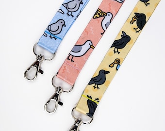 May include: Three colorful lanyards with metal clasps. The lanyards feature repeating patterns of cartoon birds. One is light blue with gray pigeons, another is pink with white seagulls, and the third is yellow with black crows.