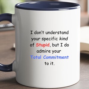 May include: White ceramic mug with a navy blue handle and interior. The mug features the text "I don't understand your specific kind of Stupid, but I do admire your Total Commitment to it." in black, red, and blue.