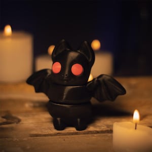 以下が含まれることがあります： 赤い目と翼を広げた黒いコウモリの置物。コウモリは木製の表面にあり、背景にはろうそくが灯っています。ハロウィーンの装飾です。