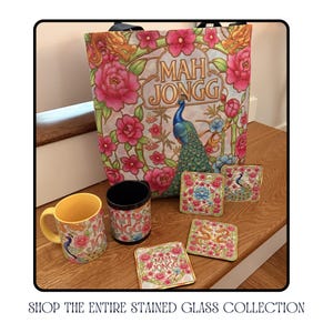 A collection of coordinating Mah Jongg-themed items, including a tote bag, mug, coasters, and a tumbler. The tote bag features a peacock and floral design with the words "Mah Jongg". The mug and coasters also have peacock and floral designs.