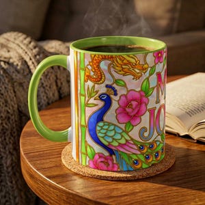 A ceramic mug with a green handle and interior, featuring a colorful peacock, dragon, and floral design. The mug has the phrase "Mah Jongg" printed on it but only the M and J are visible from this camera angle. The mug sits on a cork coaster.