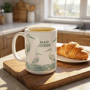 15 ounce ceramic mug with steaming liquid that resembles coffee sits on a kitchen counter on a wood cutting board and next to a plated coissant. The coffee mug shows a muted sage green design that shows the words mah Kong and four different sized herons standing on what appear to be a walkway of sea glass mosaic tiles.