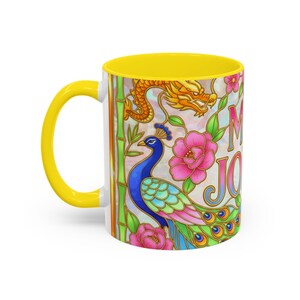 A ceramic mug with a yellow handle and interior, featuring a colorful peacock, dragon, and floral design. The mug has the phrase "Mah Jongg" printed on it but only the M and J are visible from this camera angle.