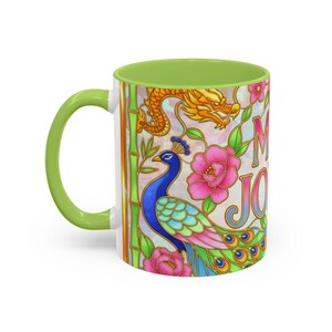 A ceramic mug with a green handle and interior, featuring a colorful peacock, dragon, and floral design. The mug has the phrase "Mah Jongg" printed on it but only the M and J are visible from this camera angle.