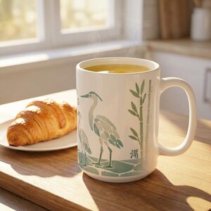 15 oz Ceramic Mug with steaming liquid presumably coffee or tea sitting on a wooden cutting board in front of a kitchen window and accompanied by a coissant. The coffee mug shows a heron and some bamboo stalks and appears to be made of sea glass mosaics in a muted sage green aesthetic