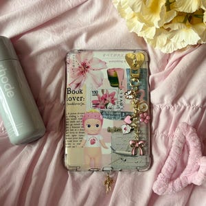 May include: Clear phone case with a collage design featuring a doll, flowers, and text. The case has a gold charm and a pink bow. The background is a pink fabric with a bottle and a flower arrangement.