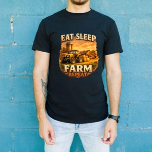 May include: A black short-sleeved t-shirt featuring a graphic print of a yellow tractor in a field with hay bales at sunset. The text EAT SLEEP FARM REPEAT is printed in a bold, distressed font above and below the central farm scene illustration.
