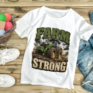 May include: A white short-sleeved t-shirt featuring a graphic print of a green tractor in a muddy field. The text "FARM" appears in green above the tractor, and the word "STRONG" appears in a textured beige font below the tractor illustration.