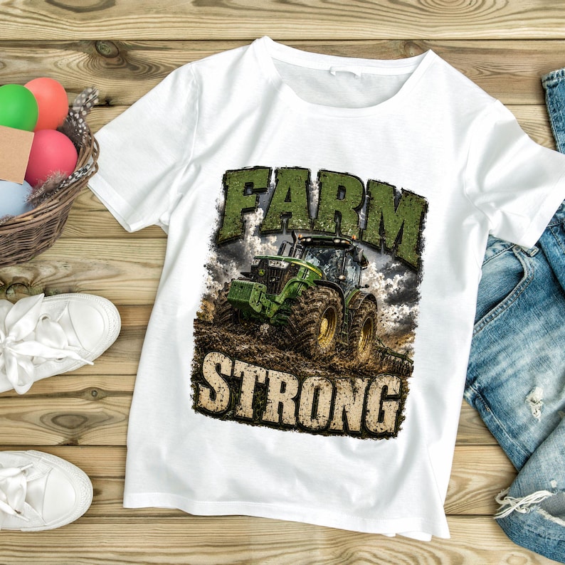 May include: A white short-sleeved t-shirt featuring a graphic print of a green tractor in a muddy field. The text "FARM" appears in green above the tractor, and the word "STRONG" appears in a textured beige font below the tractor illustration.