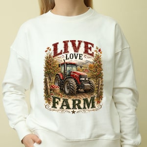 May include: A white crewneck sweatshirt featuring a graphic print of a red tractor in an orchard with a basket of apples. The text "LIVE LOVE FARM" is printed in red and green lettering above and below the tractor illustration.