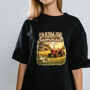 May include: A black t-shirt featuring a graphic print of a red tractor in a golden wheat field next to a wooden barn. The text above the scene reads FARM LIFE IS MY HAPPY PLACE in a rustic font. The design includes white daisies in the foreground.