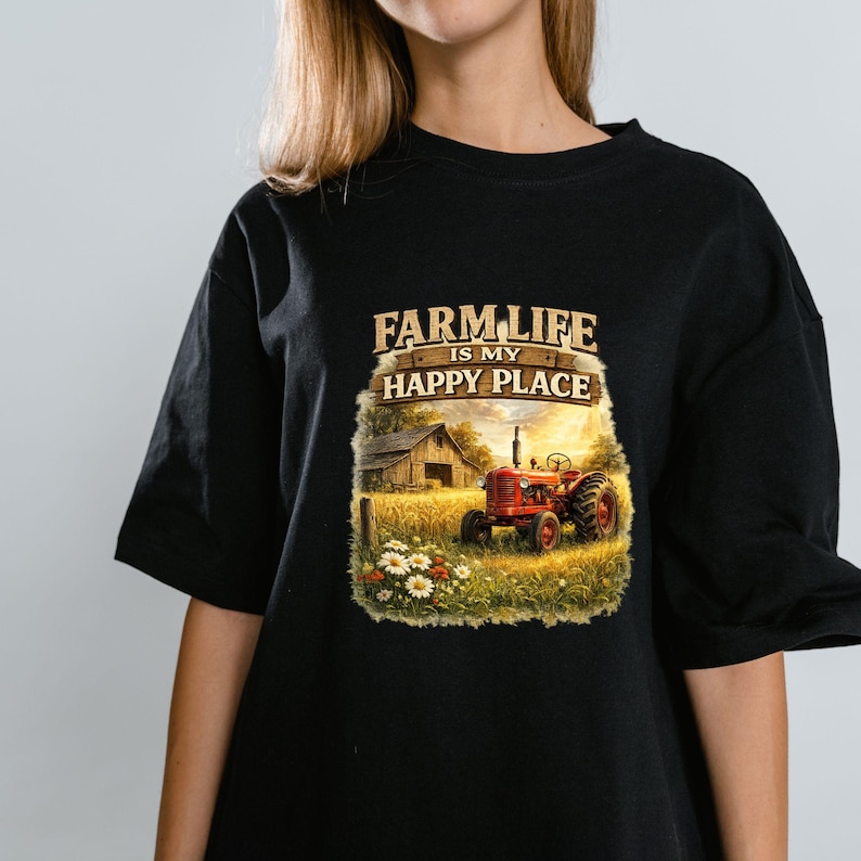 May include: A black t-shirt featuring a graphic print of a red tractor in a golden wheat field next to a wooden barn. The text above the scene reads FARM LIFE IS MY HAPPY PLACE in a rustic font. The design includes white daisies in the foreground.