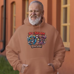 May include: A tan hoodie with a graphic design featuring the words "Six Seven" above the numbers 6 and 7, each with cartoon faces. Below the numbers is the word "BRUH". The hoodie has a hood and a front pocket.
