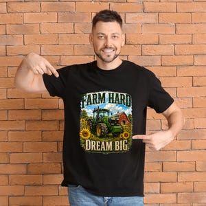 May include: A black short-sleeved t-shirt featuring a graphic print of a green tractor in a field with sunflowers and a red barn. The text above the tractor reads FARM HARD and the text below the tractor reads DREAM BIG in bold lettering.