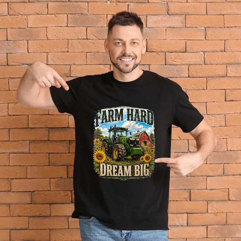 May include: A black short-sleeved t-shirt featuring a graphic print of a green tractor in a field with sunflowers and a red barn. The text above the tractor reads FARM HARD and the text below the tractor reads DREAM BIG in bold lettering.
