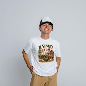 May include: A white t-shirt featuring a graphic print of a green tractor, a red barn, and chickens with the text "RAISED ON A FARM" in dark green and red lettering, paired with a white and black trucker hat and tan pants.