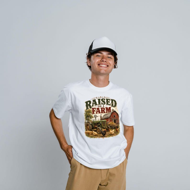 May include: A white t-shirt featuring a graphic print of a green tractor, a red barn, and chickens with the text "RAISED ON A FARM" in dark green and red lettering, paired with a white and black trucker hat and tan pants.