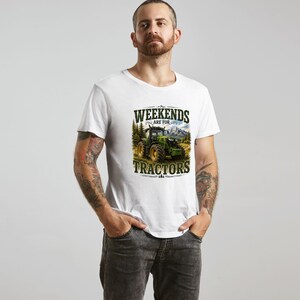 May include: A white short-sleeved t-shirt featuring a graphic print of a green tractor in a field with mountains and trees in the background. The text "WEEKENDS ARE FOR TRACTORS" is printed in dark green lettering above and below the tractor image.