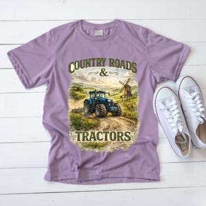 May include: A light purple short-sleeve t-shirt featuring a graphic print of a blue tractor on a dirt path with a windmill in the background. The text "COUNTRY ROADS & TRACTORS" is printed in dark green lettering above and below the illustration.