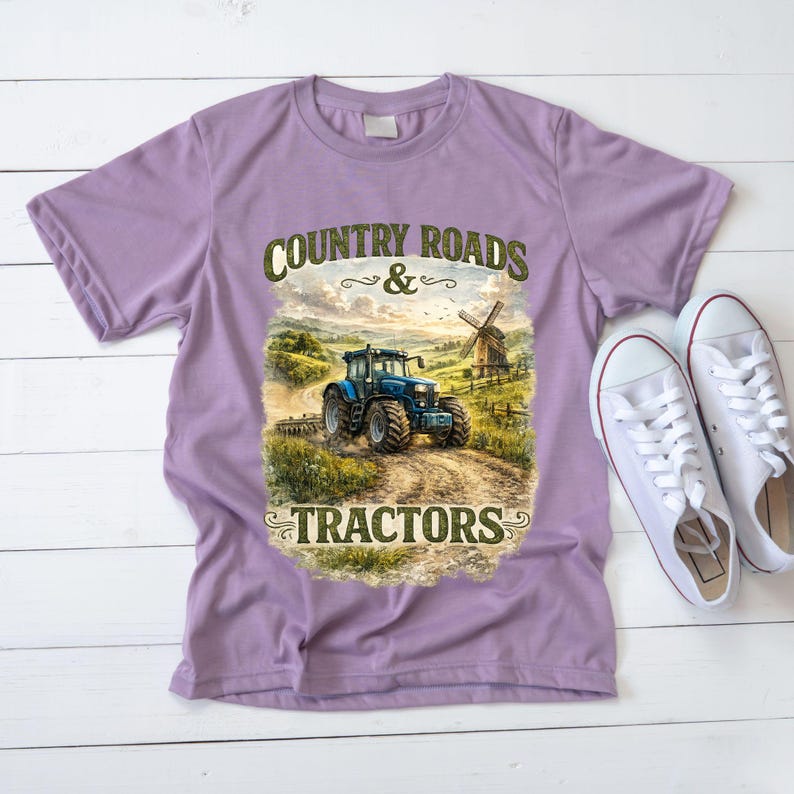 May include: A light purple short-sleeve t-shirt featuring a graphic print of a blue tractor on a dirt path with a windmill in the background. The text "COUNTRY ROADS & TRACTORS" is printed in dark green lettering above and below the illustration.