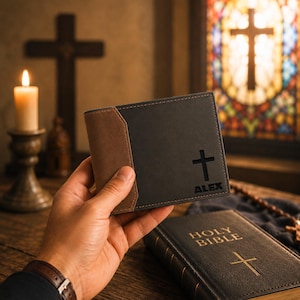 May include: A bi-fold wallet featuring a dark charcoal gray main body with a brown leather accent panel on the left side. The wallet includes a cutout cross symbol and the name ALEX embossed in black on the lower right corner.