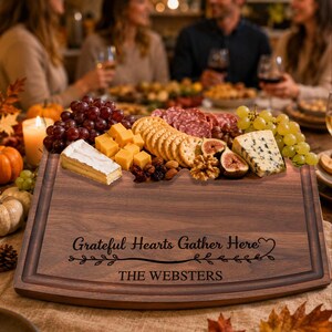 May include: A dark wood serving board featuring a carved border and the engraved text "Grateful Hearts Gather Here" above "THE WEBSTERS". The board is topped with an assortment of red grapes, cubed cheddar cheese, crackers, salami, blue cheese, and walnuts.