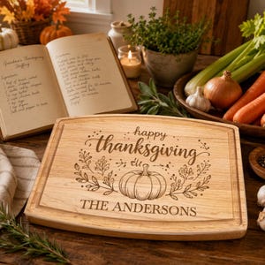 May include: A rectangular wooden cutting board featuring an engraved pumpkin illustration, floral accents, and the text "happy thanksgiving" above "THE ANDERSONS". The board has a grooved border and a light brown natural wood grain finish.