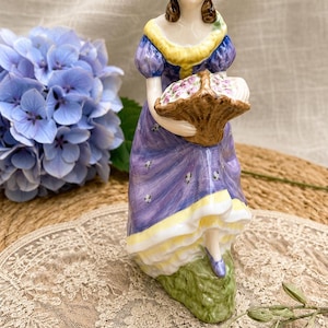 May include: A porcelain figurine of a person wearing a yellow hat and a purple dress with yellow trim, holding a brown basket filled with small pink flowers. The figure stands on a green base and features brown hair and painted facial details.