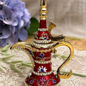 May include: A decorative red metal oil lamp with a gold-tone finish, featuring a tall spout, a curved handle, and a hinged lid. The surface is embellished with clear rhinestones, blue and green accents, and small white faux pearls at the base.