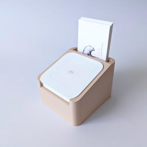 Può includere: Un organizer da scrivania beige con coperchio bianco e fessura per biglietti. I biglietti sono bianchi con un'illustrazione nera e la parola "lou" stampata sopra. L'organizer è di forma quadrata.