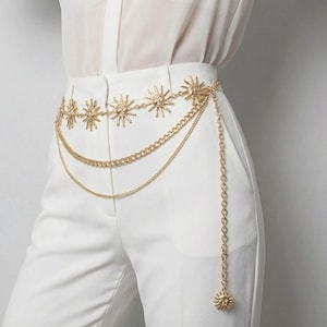 Puede incluir: Cinturón de cadena dorado con dijes en forma de sol y cadenas colgantes. El cinturón se usa sobre pantalones blancos y una camisa blanca. Los dijes de sol están espaciados uniformemente.