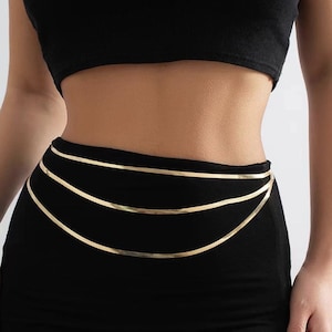 Puede incluir: Cadena de cuerpo dorada con tres finas hebras. La cadena se lleva sobre un atuendo negro, creando un efecto de capas. La joya añade un toque de elegancia y estilo al conjunto.