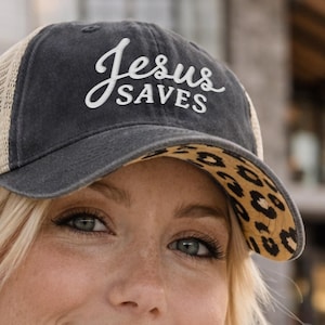Light pink women’s Christian trucker hat with white embroidered “Jesus Saves” text, two toned hat. Styled on a smiling woman with curly ponytail in front of a modern church. Vintage washed unstructured ponytail baseball cap with feminine floral details and faith-inspired boutique style.