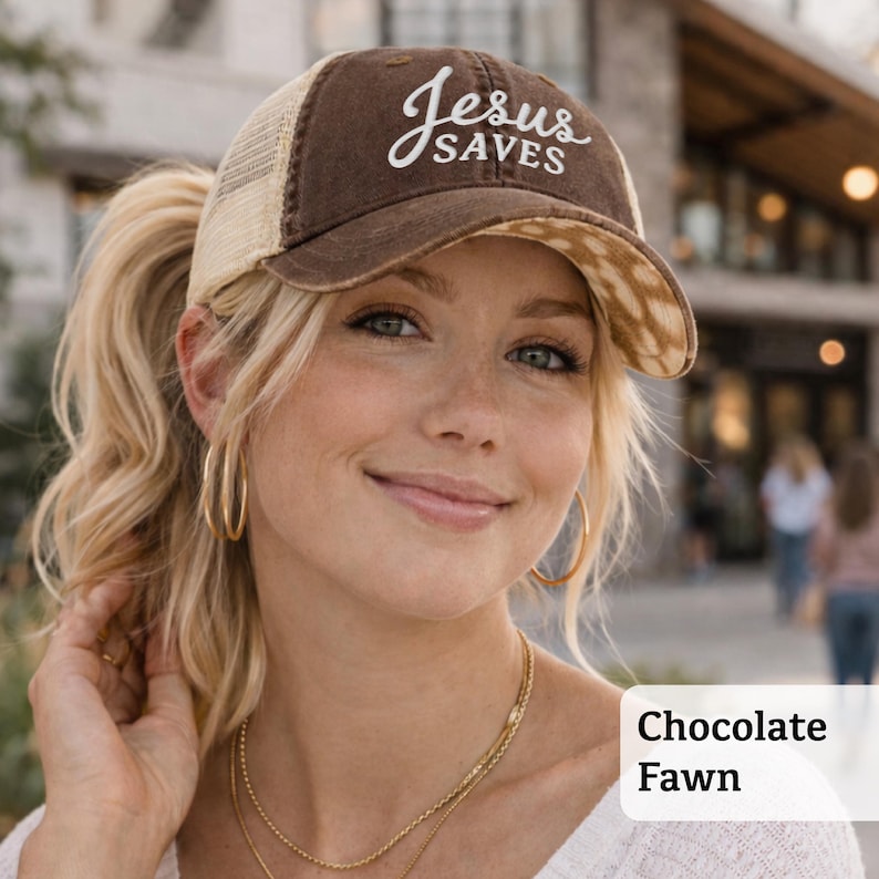 Light pink women’s Christian trucker hat with white embroidered “Jesus Saves” text, two toned hat. Styled on a smiling woman with curly ponytail in front of a modern church. Vintage washed unstructured ponytail baseball cap with feminine floral details and faith-inspired boutique style.