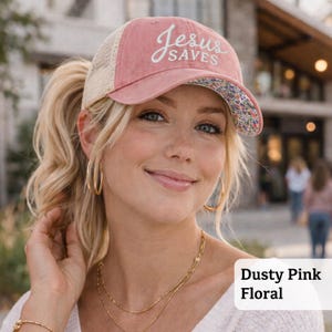 Light pink women’s Christian trucker hat with white embroidered “Jesus Saves” text, two toned hat. Styled on a smiling woman with curly ponytail in front of a modern church. Vintage washed unstructured ponytail baseball cap with feminine floral details and faith-inspired boutique style.
