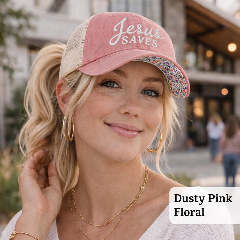 Light pink women’s Christian trucker hat with white embroidered “Jesus Saves” text, two toned hat. Styled on a smiling woman with curly ponytail in front of a modern church. Vintage washed unstructured ponytail baseball cap with feminine floral details and faith-inspired boutique style.