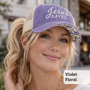 Light pink women’s Christian trucker hat with white embroidered “Jesus Saves” text, two toned hat. Styled on a smiling woman with curly ponytail in front of a modern church. Vintage washed unstructured ponytail baseball cap with feminine floral details and faith-inspired boutique style.
