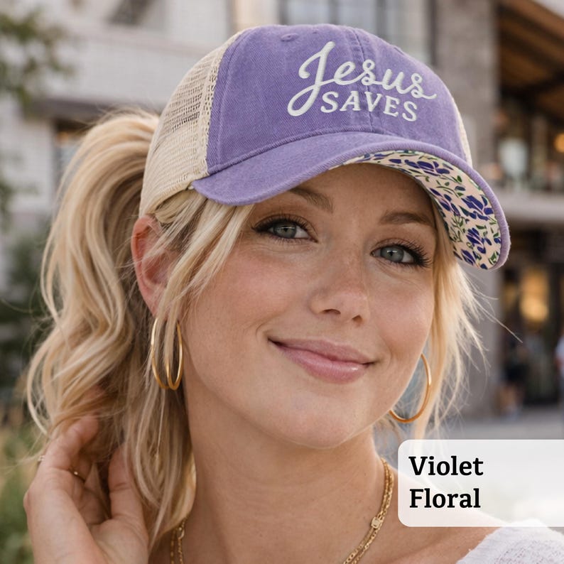 Light pink women’s Christian trucker hat with white embroidered “Jesus Saves” text, two toned hat. Styled on a smiling woman with curly ponytail in front of a modern church. Vintage washed unstructured ponytail baseball cap with feminine floral details and faith-inspired boutique style.