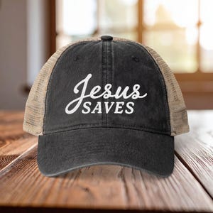 Light pink women’s Christian trucker hat with white embroidered “Jesus Saves” text, two toned hat. Styled on a smiling woman with curly ponytail in front of a modern church. Vintage washed unstructured ponytail baseball cap with feminine floral details and faith-inspired boutique style.