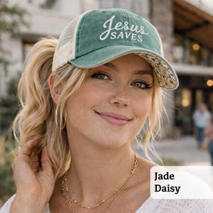Light pink women’s Christian trucker hat with white embroidered “Jesus Saves” text, two toned hat. Styled on a smiling woman with curly ponytail in front of a modern church. Vintage washed unstructured ponytail baseball cap with feminine floral details and faith-inspired boutique style.