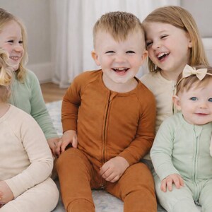 K&ouml;nnte beinhalten: Eine Gruppe von Kindern in passenden Schlafanz&uuml;gen in verschiedenen Farben, darunter Rost, Salbeigr&uuml;n und Creme. Die Kinder l&auml;cheln und lachen, was eine fr&ouml;hliche und herzerw&auml;rmende Szene ergibt. Die Schlafanz&uuml;ge haben einen Rei&szlig;verschluss.