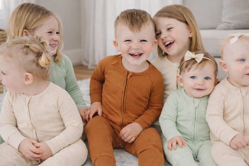 K&ouml;nnte beinhalten: Eine Gruppe von Kindern in passenden Schlafanz&uuml;gen in verschiedenen Farben, darunter Rost, Salbeigr&uuml;n und Creme. Die Kinder l&auml;cheln und lachen, was eine fr&ouml;hliche und herzerw&auml;rmende Szene ergibt. Die Schlafanz&uuml;ge haben einen Rei&szlig;verschluss.