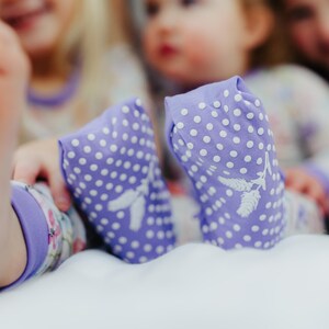 以下が含まれることがあります： 白のドットと白い花の模様が入った、ラベンダー色のフットカバーのクローズアップ。フットカバーはパジャマセットの一部です。背景には子供たちがいて、画像はソフトフォーカスです。