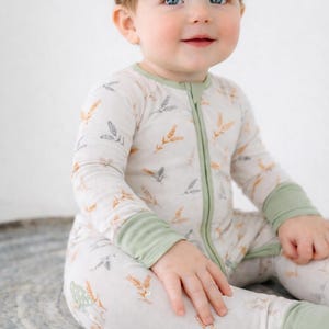 K&ouml;nnte beinhalten: Ein Baby tr&auml;gt einen hellfarbenen Strampler mit gr&uuml;nem Rei&szlig;verschluss und B&uuml;ndchen. Der Strampler hat ein Muster aus Weizen&auml;hren in Orange und Grau. Ein beiges Schleifen-Stirnband ziert den Kopf des Babys.